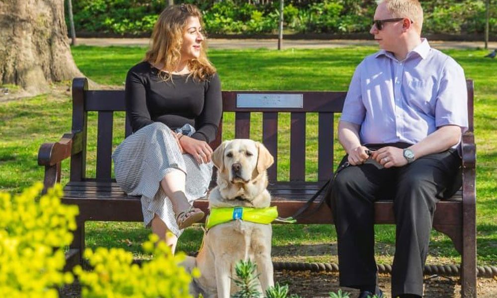 Photo of visually impaired individual in park with guide dog and friend